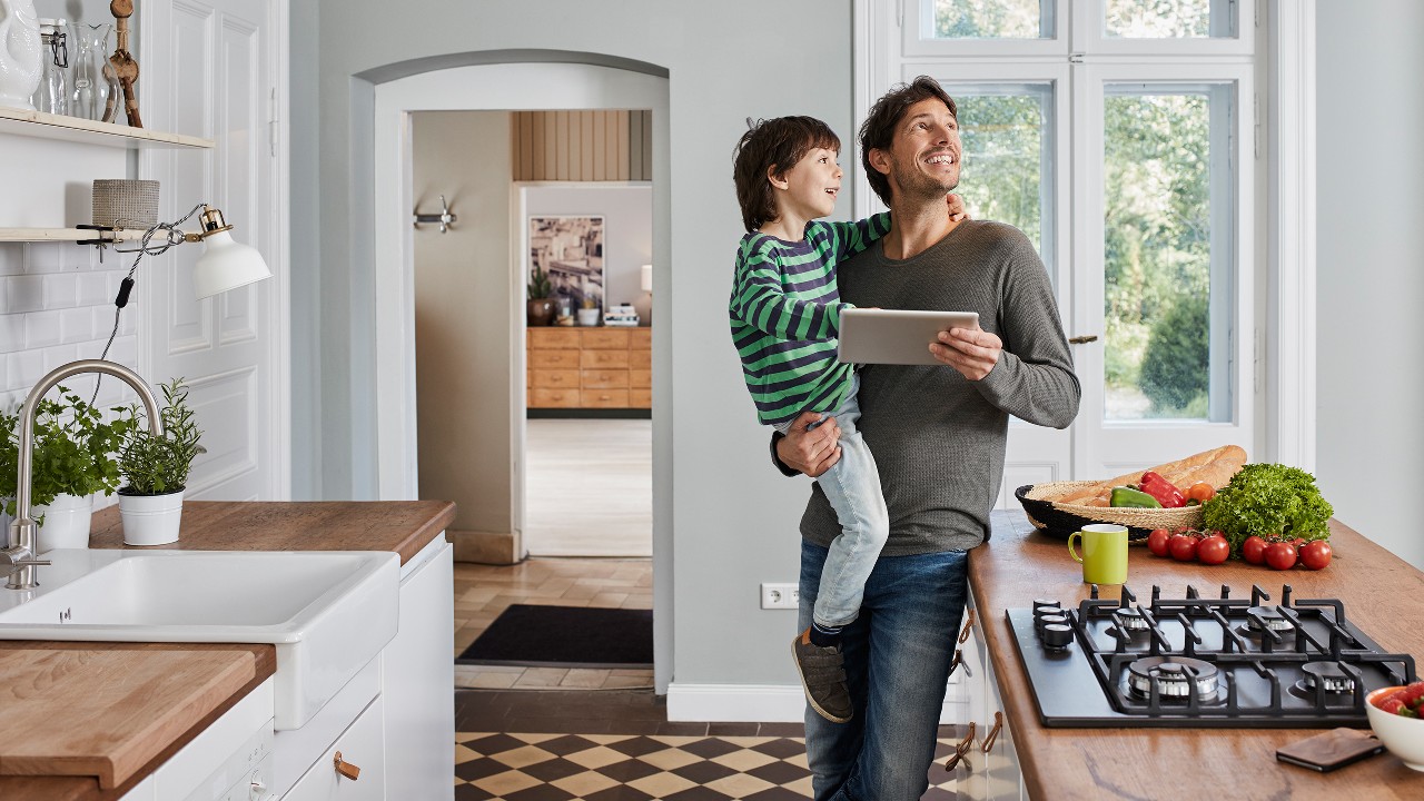 A father happily holding his son with a tablet in the other hand, looking around in a kitchen;  image used for HSBC Sri Lanka credit card household offer page